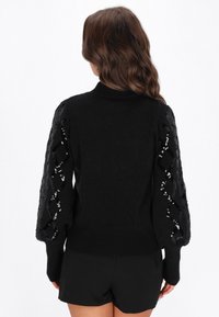 Fekete pulóver magas gallérral, texturált kötött testtel és puffos ujjal, fekete flitteres díszítéssel, gyémánt mintázatban.
