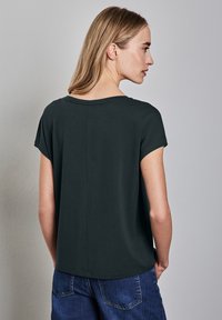 Tmavě zelené tričko s krátkým rukávem vyrobené z měkkého materiálu, s kulatým výstřihem a volným střihem, nošené s modrými denimi.