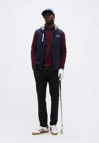 Άνδρας με μπλε καπέλο, ναυτικό γιλέκο, μαροκινό πουλόβερ, μαύρο παντελόνι και λευκό γάντι του γκολφ κρατάει ένα ρόπαλο γκολφ, όρθιος μπροστά σε λευκό φόντο.