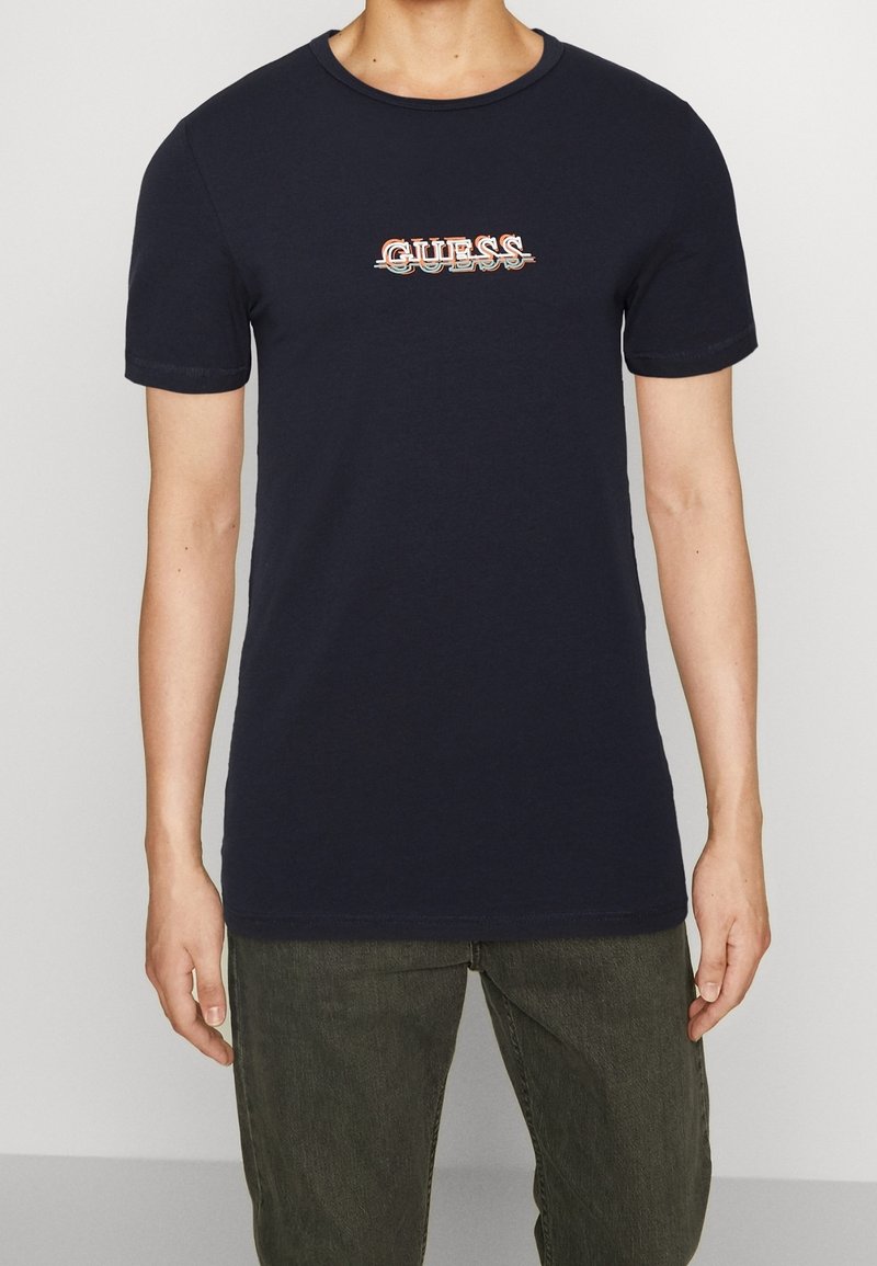 Navyblaue Baumwoll-T-Shirt mit kurzen Ärmeln, Rundhalskragen und einem Vintage-Style "GUESS" Logo in mehreren Farben über der Brust.