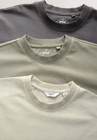Trois t-shirts à col rond pliés en gris foncé, vert olive et beige, chacun avec des étiquettes de marque visibles portant la mention Next.