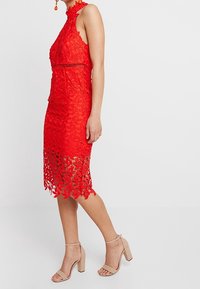 Femme portant une robe midi en dentelle rouge sans manches avec des découpes florales et des sandales à talons blocs beige à bride de cheville, se tenant devant un fond blanc uni.