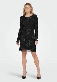 Czarna sukienka z długim rękawem z kwiatowym haftem, miękka w dotyku, o długości do kolan, ze przezroczystymi akcentami. Do kompletu czarne szpilki na spiczastym nosku.