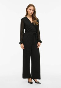 Črn kombinezon z dolgimi, prosojnimi, pikčastimi rokavi, ovitjem, zavezanim pasom in širokimi hlačami. Združeno s črnimi čevlji s koničastim nosom in zlatenimi detajli.