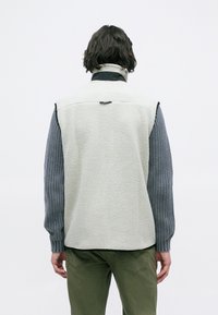 Pessoa a usar um colete de fleece texturizado branco, um suéter de malha cinza de mangas longas e calças verde-oliva, visto de costas contra um fundo simples.
