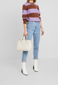 Vit läderhandväska med dubbla handtag, slät yta. Modellen bär en lila och brun randig tröja, ljusblå jeans och vita ankelstövlar.