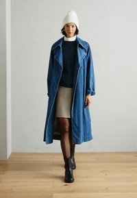 Gabardina de mezclilla con solapas amplias, dos botones frontales, superpuesta sobre un suéter oscuro y una falda corta, combinada con botas de tobillo negras.