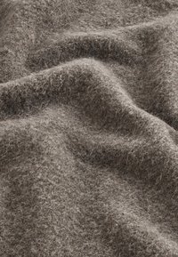 Weiches, strukturiertes braunes Gewebe mit einem flauschigen Aussehen, das eine Mischung aus hellen und dunklen Fasern in einem drapierten, wellenförmigen Muster zeigt.