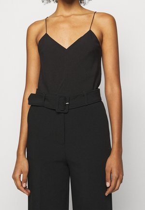 Kobieta ubrana w czarny bezrękawnik z dekoltem w serek i cienkimi ramiączkami, włożony do wysokich czarnych spodni z szerokim paskiem.