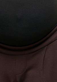 Gros plan d'une personne portant un haut noir rentré dans un pantalon taille haute marron foncé avec des coutures visibles et une coupe ajustée.