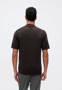 Brun kortermet polo skjorte med krage, ribbestrikket tekstur og rett søm, sett fra baksiden, iført grå bukser.