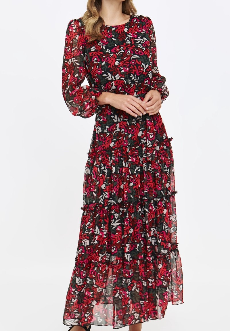 Femme portant une longue robe noire transparente avec un imprimé floral rouge et blanc et de longues manches bouffantes, debout les mains jointes à la taille.