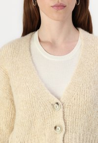Cardigan beige, texture tricotée, design en V, comportant deux grands boutons. Porté par-dessus un haut crème clair à côtes.