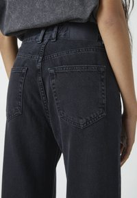 Lähikuva henkilöstä, joka käyttää korkeavyötäröisiä tummansinisistä farkuista valmistettuja farkkuja, joissa on takataskut, sekä sisätaitettuna tummanharmaata puseroa.