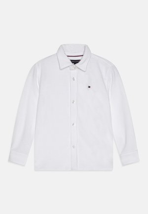 Λευκό πουκάμισο πόλο με μακριά μανίκια, κουμπιά μπροστά, ριγωτή υφή και ένα μικρό κεντημένο λογότυπο στο στήθος. Κλασικός σχεδιασμός γιακά.