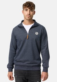 Marineblå quarter-zip pullover lavet af tekstureret stof, med lynlås og et emblem på venstre bryst. Ribbestrikkede mansjetter og kant.
