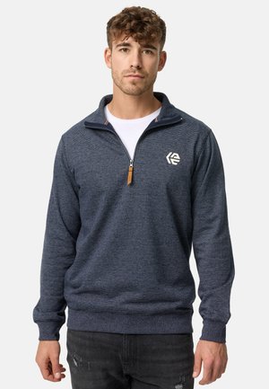 Marineblå quarter-zip pullover lavet af tekstureret stof, med lynlås og et emblem på venstre bryst. Ribbestrikkede mansjetter og kant.