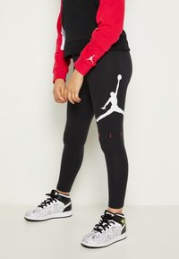 Czarne legginsy z dużym białym logo Jumpman na lewym udzie, zestawione z czarnym i czerwonym krótkim hoodie oraz wzorzystymi trampkami.