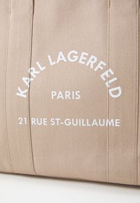 Borsa tote beige realizzata in tessuto testurizzato, con testo stampato bianco: "KARL LAGERFELD," "PARIS," e "21 RUE ST-GUILLAUME." Cuciture a vista.