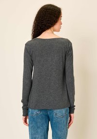 Moteris su garbanotais plaukais, vilkinti tamsiai pilką ilgomis rankovėmis marškinėlį ir mėlynas džinsas, žiūrinti į nugarą prieš paprastą smėlio spalvos foną.