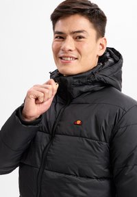 Schwarze Winterjacke mit einem Reißverschluss vorne und verstellbarem Kapuzen. Auffälliges, gesticktes Logo auf der Brust. Glatte, glänzende Textur.