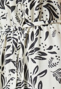 Motif de feuilles noires et de points sur fond crème. Tissu doux avec une taille froncée et un détail de ceinture nouée. Texture visible et variation de design.