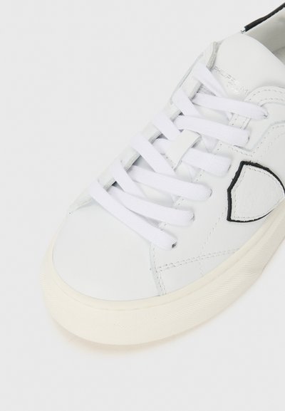 Białe skórzane sneakersy z płaskimi białymi sznurowadłami. Posiadają teksturowaną boczną część oraz okrągły czarny akcent. Guma podeszwy ma gładkie wykończenie.