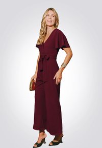 Burgundský overal s krátkými rozšířenými rukávy, svázaným pasem, širokými nohavicemi a hladkou texturou. V kombinaci s malou kabelkou s zlatými akcenty.