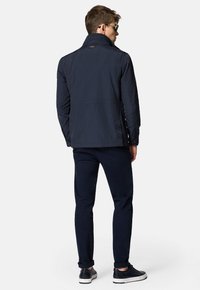 Granatowa kurtka przeciwwiatrowa z wysokim kołnierzem, zamykanymi kieszeniami bocznymi i teksturowanym wykończeniem. Noszona z ciemnymi spodniami i stylowymi sneakersami.