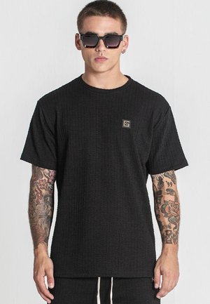 Camiseta negra de textura con mangas cortas, cuello redondo y un pequeño parche con el logo. Se lleva con gafas de sol oscuras.
