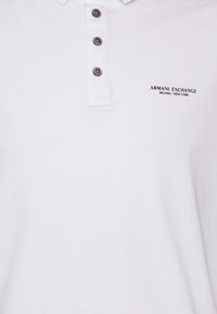 Vitt polo-shirt med tre mörka knappar, mjukt bomullsmaterial. Har en liten svart logotyp som säger "ARMANI EXCHANGE MILANO / NEW YORK."