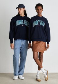 Sudaderas navy con el texto "TOMMY JEANS", detalles en azul claro, combinadas con jeans azul claro y una falda de cuero marrón. Zapatillas blancas con detalles grises.