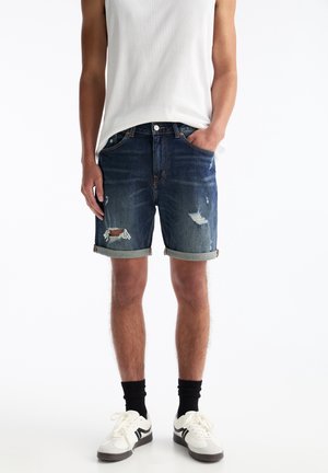 Dunkelblaue Jeansshorts mit ausgefransten Rissen und umgeschlagenen Säumen. Kombiniert mit einem hellen Tanktop und Sneakers mit schwarz-weißen Akzenten.
