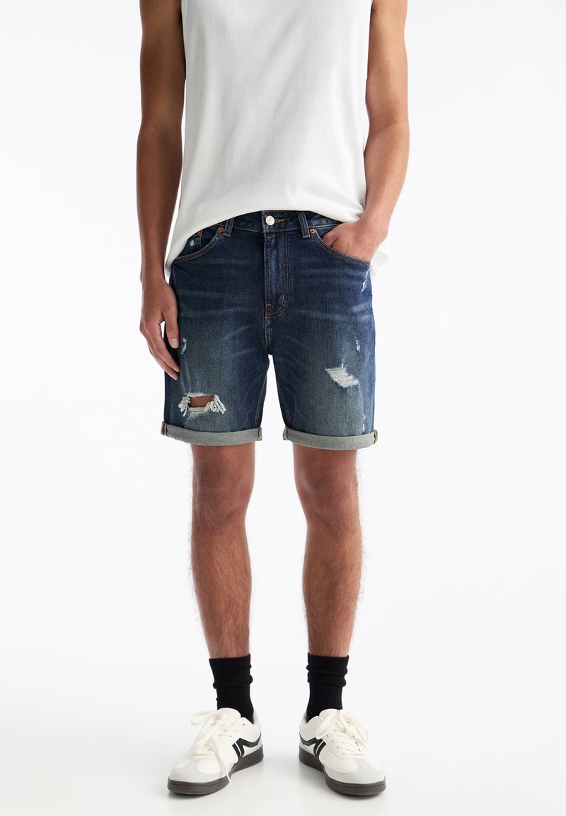 Shorts en jean bleu foncé avec des déchirures usées et des revers roulés. Associés à un débardeur clair et des baskets avec des accents noirs et blancs.