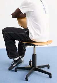 Trä svängbar stol med rund sits, svart metallbas, justerbar höjd, och en person klädd i en vit t-shirt och m örka byxor som sitter på den.