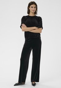 Top in velluto nero con maniche corte e a palloncino, abbinato a pantaloni in velluto neri a gamba larga. La vestibilità slim e la texture liscia creano un aspetto coerente.