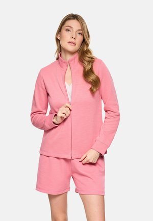 Mujer joven con el cabello largo y ondulado, vistiendo una chaqueta rosa con cremallera y pantalones cortos a juego, posando frente a un fondo claro y liso.