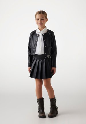 Schwarze Kunstlederjacke mit Druckknöpfen über einem weißen Hemd. Schwarzer plissierter Rock und braune kniehohe Stiefel mit Schnallen.