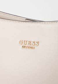 Borsa in ecopelle color crema con superficie testurizzata, caratterizzata dal logo dorato "GUESS EST 1981" e cuciture nere sui bordi.