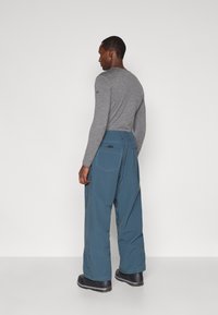 Muškarac u sivoj majici s dugim rukavima i širokim plavim hlačama s velikim stražnjim džepovima, stoji na jednostavnoj bijeloj pozadini, okrenut bočno.