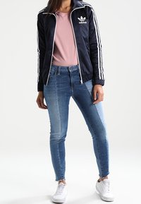 Chaqueta deportiva azul marino con rayas blancas y logo, camiseta rosa, jeans ajustados de lavado claro y zapatillas blancas. Atuendo atlético casual.