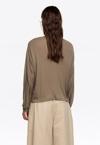 Bež majica s dugim rukavima opuštenog kroja, s rebrastom teksturom i zaobljenim rubom. Uparena sa svijetlim širokim hlačama.