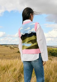 Frau mit langen dunklen Haaren, die eine mit Landschaftsmuster verzierte Wollweste, ein weißes Hemd und Jeans trägt, steht auf einer Wiese unter einem bewölkten Himmel.
