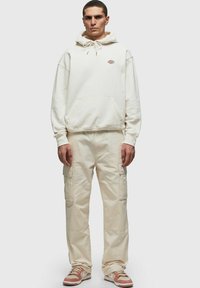 Sudadera blanca con cordones, bolsillo canguro, combinada con pantalones de carga de color beige claro con bolsillos laterales. Llevado con zapatillas marrones y blancas.