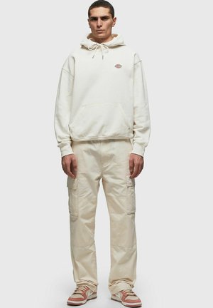 Sudadera blanca con cordones, bolsillo canguro, combinada con pantalones de carga de color beige claro con bolsillos laterales. Llevado con zapatillas marrones y blancas.