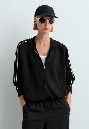 Personne portant un sweat à capuche noir zippé avec des bandes blanches sur les manches, un pantalon noir, une casquette noire et des lunettes de soleil blanches, debout les mains dans les poches.