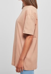 Světle růžové oversized bavlněné tričko s krátkým rukávem, kulatým výstřihem a volným střihem, nošené přes modré kalhoty. Hladká textura, ležérní design.