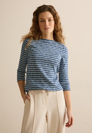 Blauw-wit gestreepte gebreide top met driekwart mouwen, bootneklijn, gestructureerde stof, gecombineerd met lichtbeige broeken.