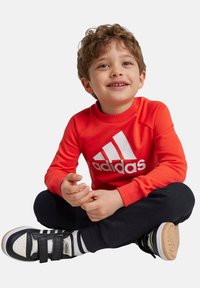 Sudadera roja de manga larga con el logo blanco de "adidas", combinada con pantalones negros y zapatos negros con rayas blancas y negras y hebillas.