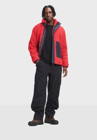 Czerwony polarowy kurta z granatowymi akcentami, z zamkiem z przodu, wysokim kołnierzem i kieszenią naszywaną. Czarne spodnie w luźnym kroju i solidne obuwie.